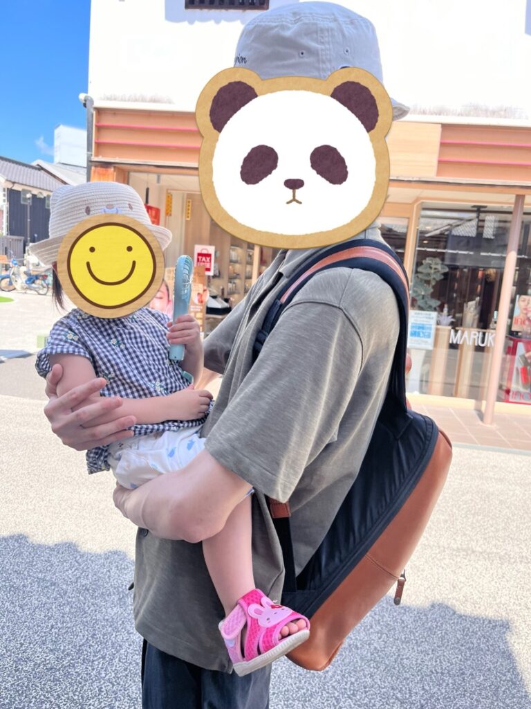 倉敷美観地区の通りで、帽子をかぶった大人が子どもを抱っこしてハンディーファンを持っている写真(顔はスタンプで隠れている)