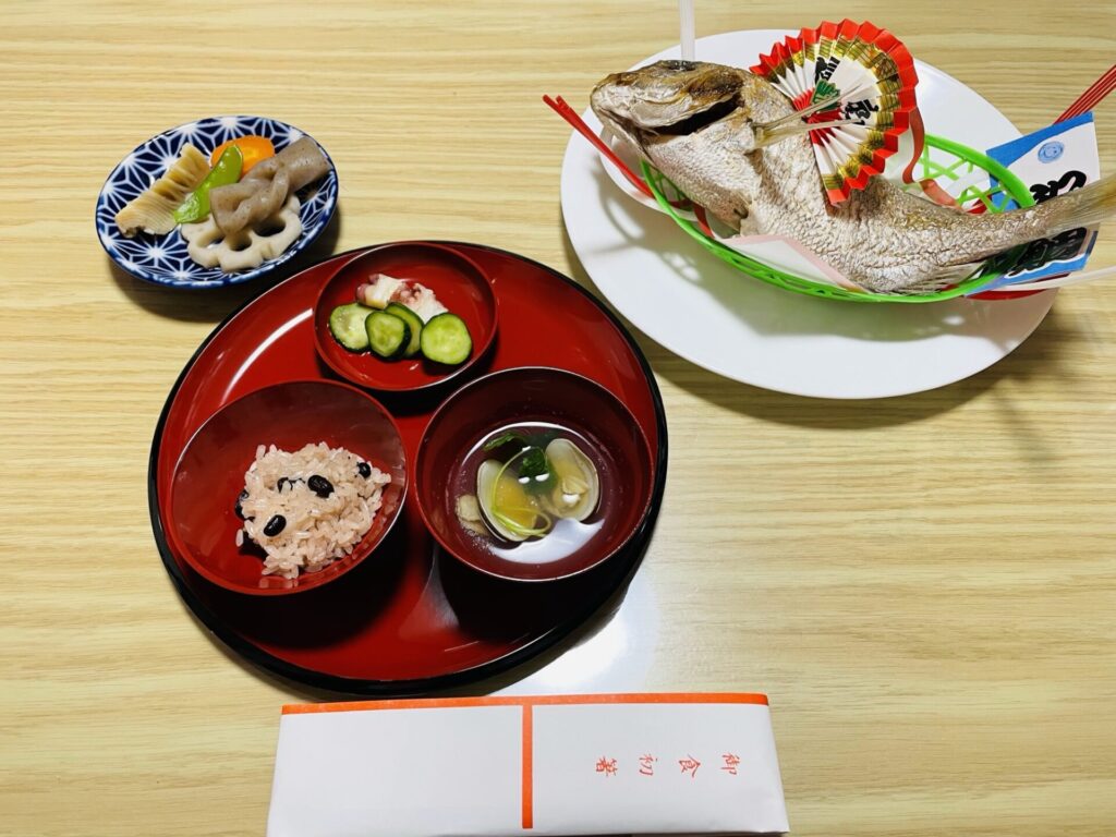 スーパーで焼いてもらった鯛と炊飯器で炊いた赤飯などを並べたお食い初め膳の写真。