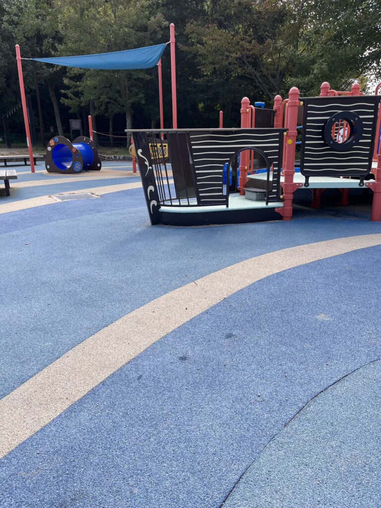 船のかたちをしたカラフルな遊具と青いゴム舗装の床が広がる広場。しあわせの村の小さい子向け遊び場