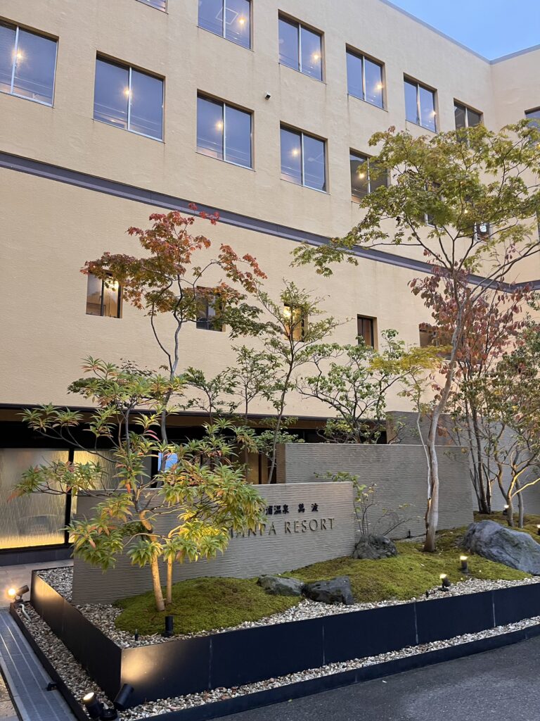 ホテル萬波の外観とエントランス。植栽とライトアップされた看板が配置されている。