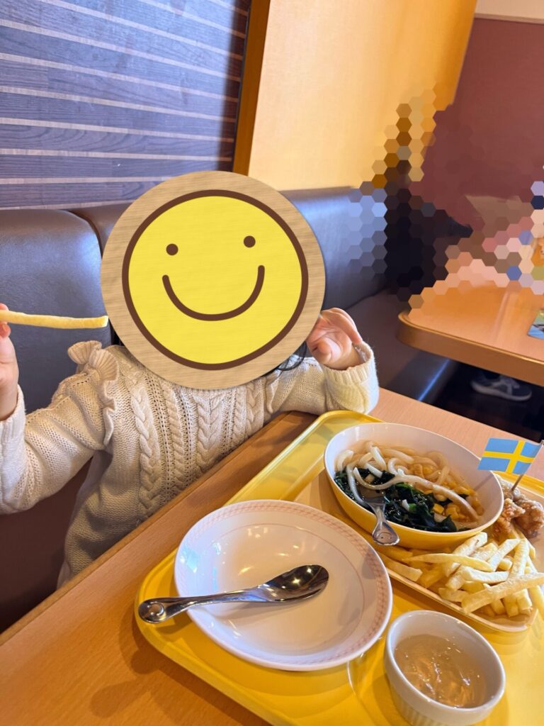 ファミレスで黄色い子ども用プレートにうどんやポテトが並び、3歳の娘が席で食事をしている様子。
