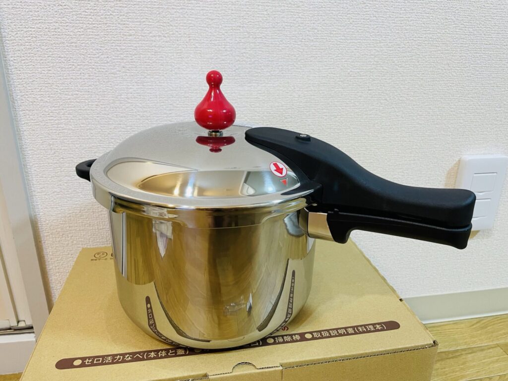 ゼロ活力鍋の本体を箱の上に置いた状態で撮影した写真。赤いおもりとステンレスの光沢が特徴的な圧力鍋が映っている。