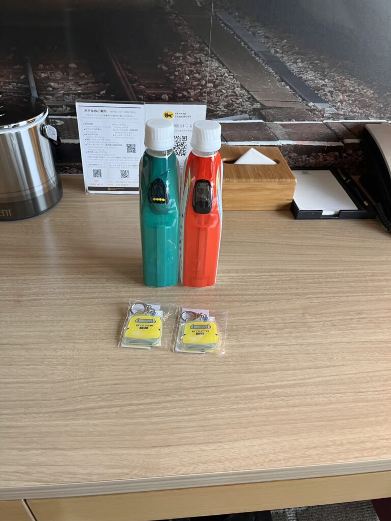 新幹線デザインのドリンクボトルとキーホルダーが並ぶホテル客室のデスク。